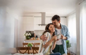 Imagem de um casal sorridente em uma cozinha iluminada, se abraçando e segurando canecas em um apartamento aconchegante, repleto de luz natural e plantas