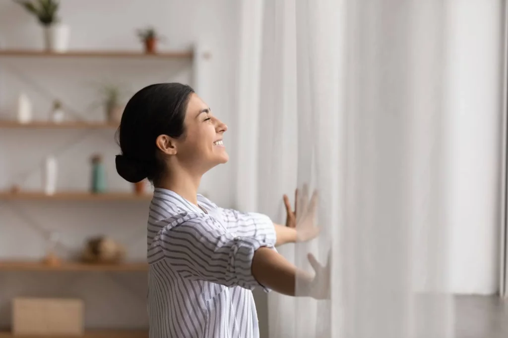 Imagem de uma mulher sorridente, vestindo uma camisa listrada, está ao lado de uma janela, abrindo suavemente as cortinas brancas. A luz do sol entra iluminando seu rosto. As plantas e a decoração estão embaçadas ao fundo