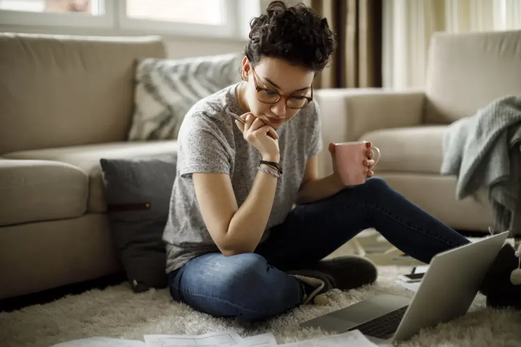 Imagem de uma mulher sentada no chão da sala segurando uma xícara de café enquanto analisa alguns documentos que estão no chão ao lado de um notebook para ilustrar matéria sobre como funciona o seguro fiança locatícia