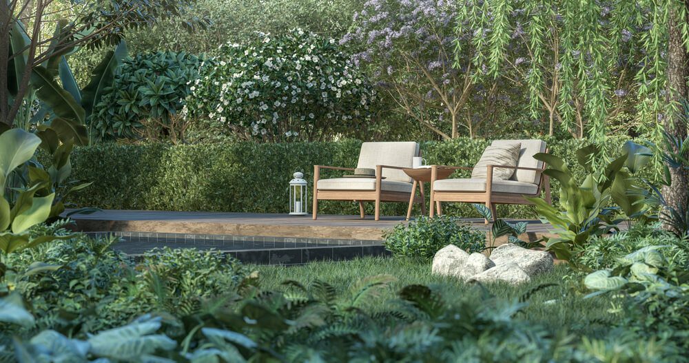 Duas cadeiras de madeira almofadadas e uma pequena mesa repousam sobre um deck de madeira em um exuberante jardim com muros simples e bonitos, cercado por plantas verdes, flores desabrochando e árvores. Uma lanterna decorativa está colocada ao lado das cadeiras.