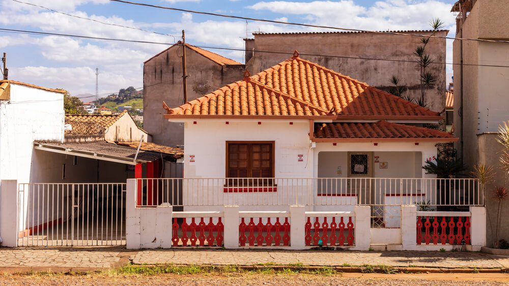 Uma pequena casa branca com telhado de telhas vermelhas e muros simples e bonitos ao longo do jardim cercado da frente fica em uma rua tranquila sob um céu parcialmente nublado, com prédios vizinhos visíveis atrás dela.