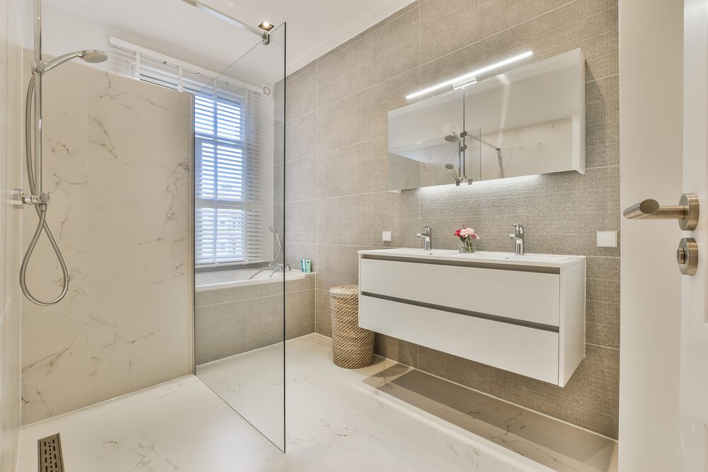 Banheiro moderno com box amplo, pia dupla flutuante, grande armário espelhado e banheira junto a janela com persianas. Tons neutros de bege e branco oferecem vibrações calmas – perfeito para quem busca ideias para banheiro elegante e funcional.