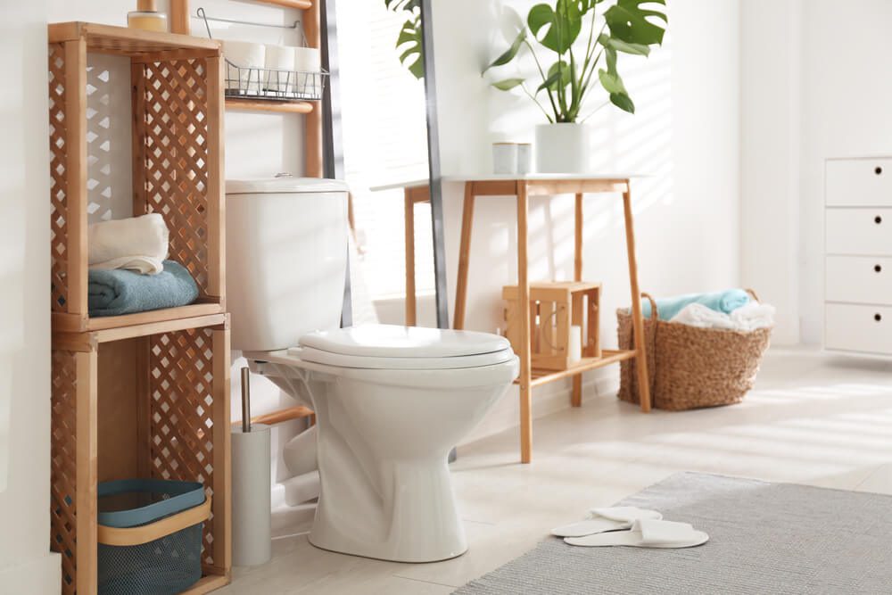 Banheiro moderno e bem iluminado com vaso sanitário branco, prateleiras de madeira, vaso de plantas, cesto de vime, toalhas, chinelos e luz solar entrando pela janela - perfeição para quem busca ideias para banheiro minimalista e organizado.