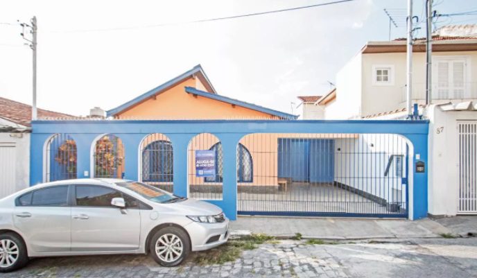 Uma pequena casa laranja com detalhes azuis e muros simples e bonitos, além de um portão azul, vista da rua. Um carro prateado está estacionado em frente, na calçada de paralelepípedos. Uma placa de "Aluga-se" é visível atrás do portão.