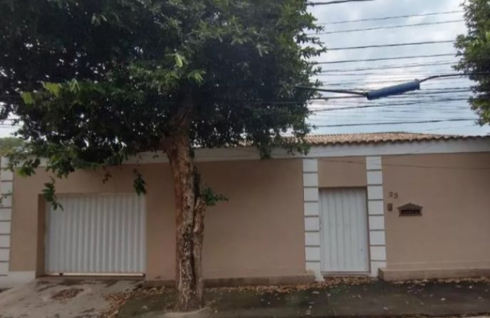Uma casa bege com muros simples e bonitos, uma porta de garagem e portão brancos, parcialmente sombreada por uma árvore. Acima, várias linhas de energia cruzam o céu, com um dispositivo cilíndrico azul preso a elas.