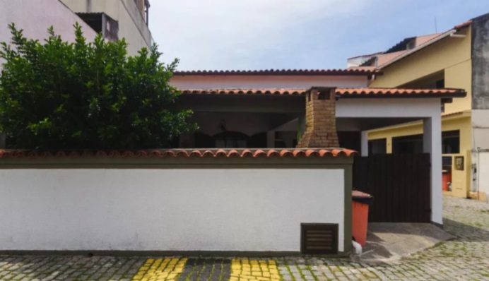 Casa térrea com telhado de telhas vermelhas, fachada branca e muros simples e bonitos na frente. À esquerda, há um arbusto, com chaminé, portão preto e calçada de paralelepípedos com marcações de tinta amarela.