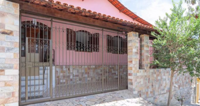 Uma casa com exterior rosa, muros simples e bonitos de telhas de pedra nas paredes inferiores e um telhado de telhas marrons. A fachada apresenta portões de ferro forjado, uma passarela de pedra e uma pequena árvore à direita.