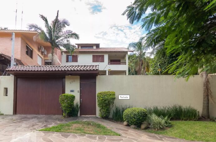 Uma casa de dois andares com telhado de telhas vermelhas, paredes bege e muros simples e bonitos. Vegetação exuberante, palmeiras e arbustos cercam a entrada, e uma placa branca com a inscrição Fabiana adorna a parede frontal.