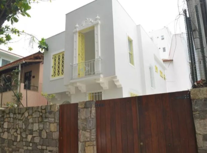 Casa branca de dois andares com janelas com detalhes amarelos e uma pequena varanda, parcialmente escondida por muros simples e bonitos de pedra e madeira, situada entre outras casas e árvores.