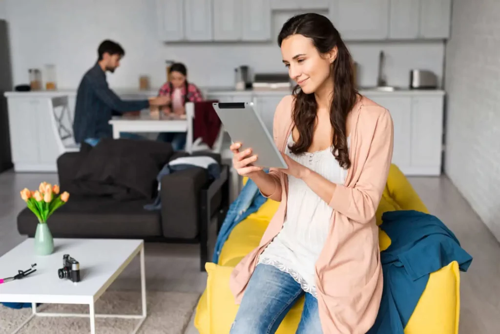 Imagem de Uma mulher sentada em um sofá amarelo usando um tablet para ilustrar matéria sobre o valor da consultoria imobiliária. Ao fundo, um homem e uma mulher conversam em uma mesa em uma sala de estar moderna e iluminada, com tulipas e uma câmera na mesa de centro.