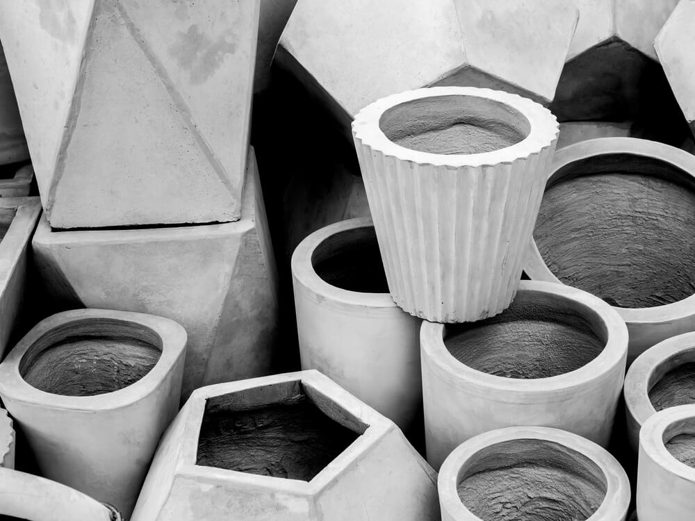 Uma foto em preto e branco mostrando uma coleção de vasos de cimento vazios, de formas geométricas, empilhados, destacando suas variadas texturas e formas.