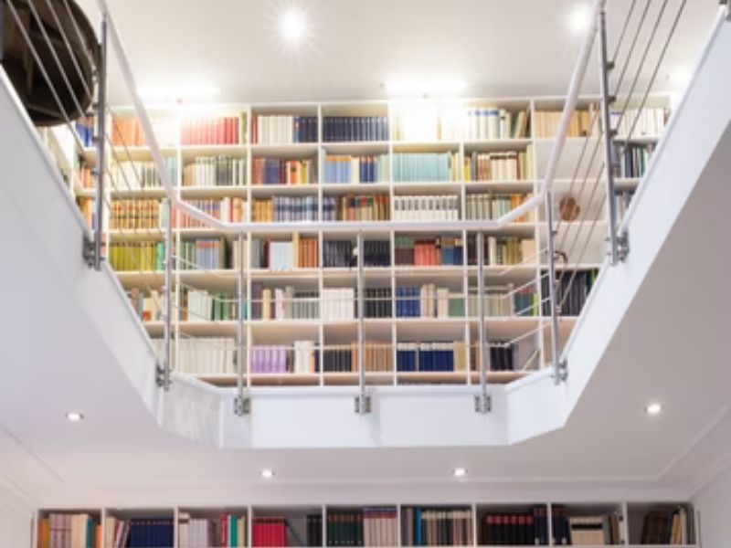 Uma vista de estantes altas e brancas repletas de livros coloridos, vistas de um andar inferior através de uma escada moderna com corrimãos de metal e luzes brilhantes no teto — uma inspiração perfeita para sua própria biblioteca em casa.
