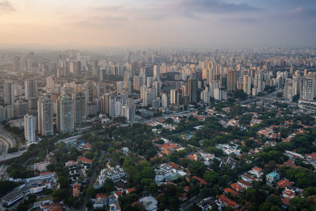 Imagem da vista aérea do Brooklin, em São Paulo, mostra aglomerados de arranha-céus altos ao longe e uma área residencial com casas e árvores em primeiro plano ao pôr do sol.