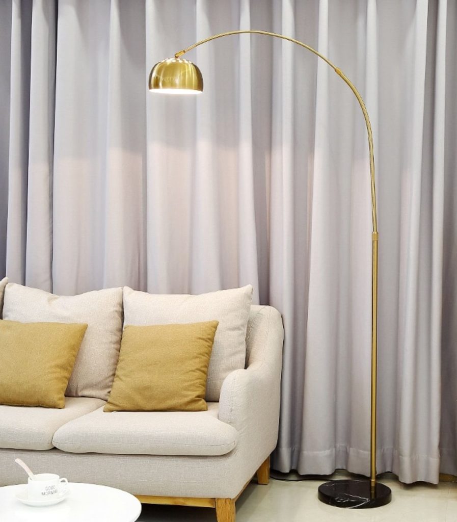 Uma sala de estar moderna com um sofá bege com duas almofadas amarelo-mostarda, uma luminária de chão dourada com cúpula, cortinas cinza-claro e elegantes objetos de decoração para sala completando o visual.