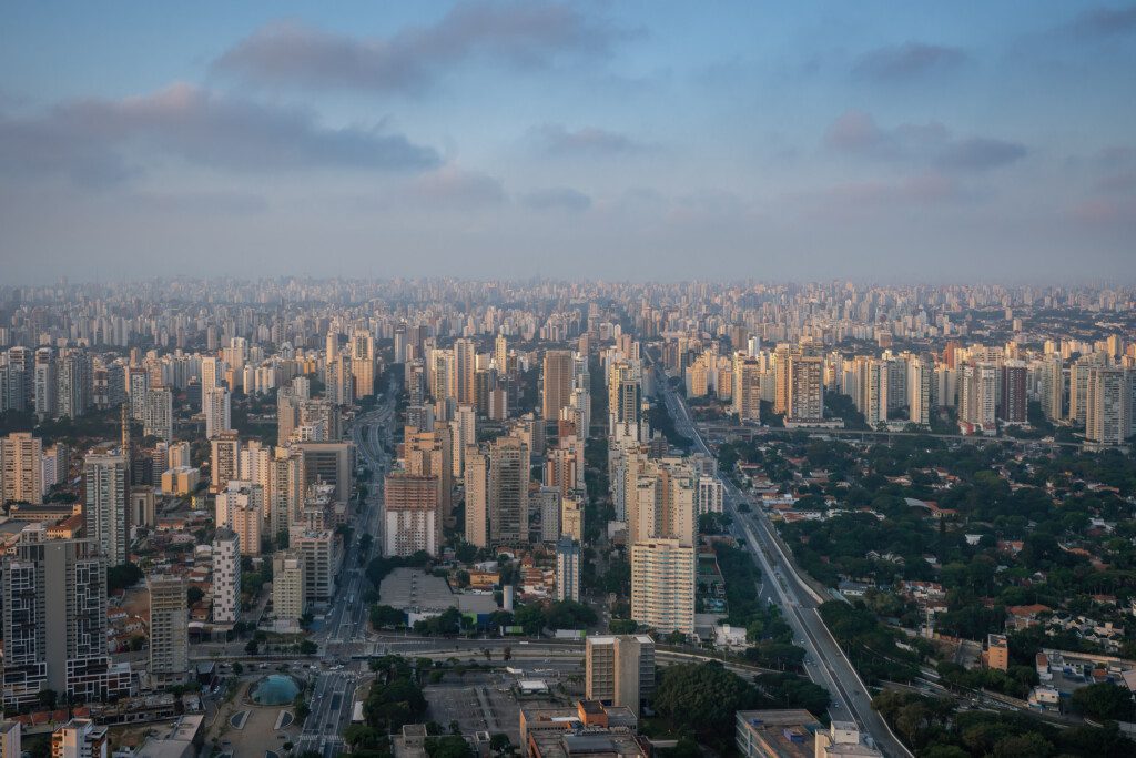Imagem da vista aérea do bairro Santo Amaro, em São Paulo, mostra uma paisagem urbana extensa com muitos prédios altos, ruas arborizadas e uma estrada principal que atravessa o centro sob um céu parcialmente nublado.