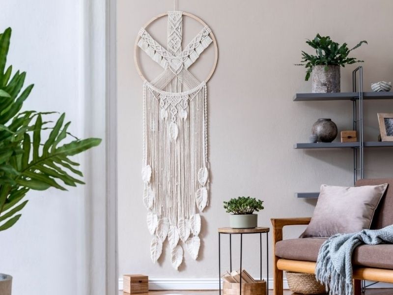 Uma sala de estar aconchegante no estilo Japandi, com uma parede bege com uma grande tapeçaria de macramê, uma cadeira de madeira com almofadas cinza, plantas verdes e prateleiras modernas adornadas com itens decorativos.
