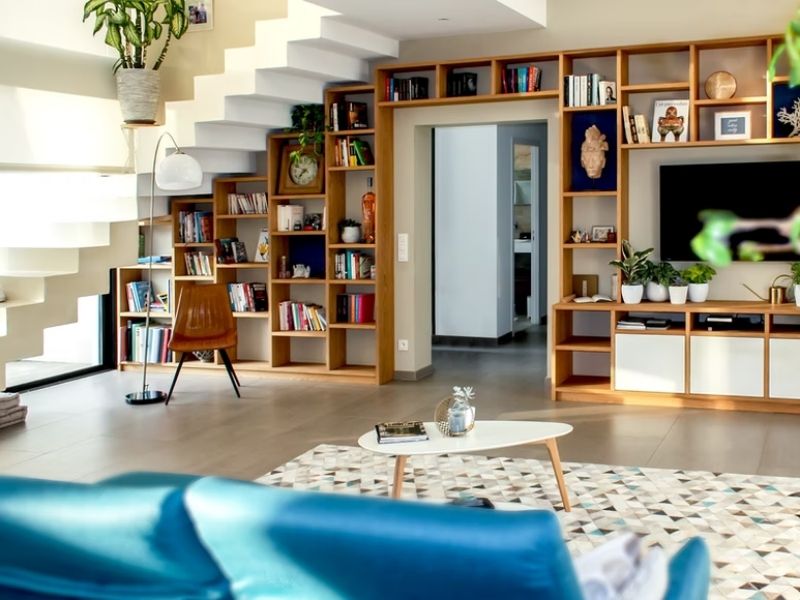 Uma sala de estar moderna e iluminada com prateleiras de madeira embutidas que criam uma sensação aconchegante de biblioteca em casa, uma TV, livros, plantas, uma escada em zigue-zague, cadeira de madeira, tapete estampado e uma mesa de centro com uma pequena planta e um livro sobre ela.