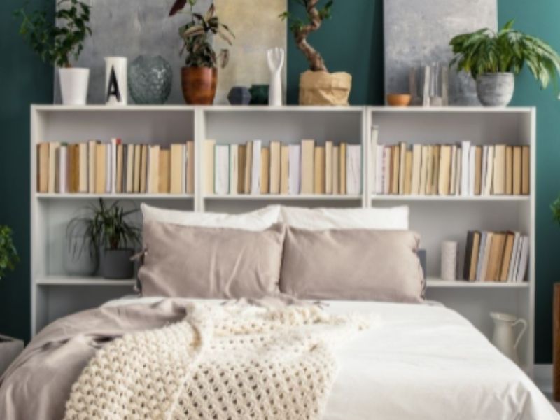 Um quarto aconchegante com uma cama impecavelmente arrumada em frente a uma biblioteca em casa branca repleta de livros e plantas. As paredes verdes e os diversos itens decorativos nas prateleiras adicionam charme, enquanto um cobertor de tricô grosso cobre a cama.