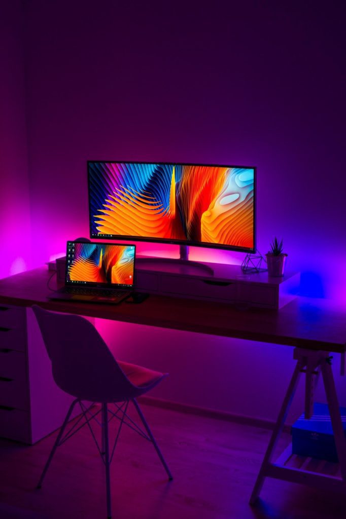 Uma mesa moderna com um monitor e um laptop exibindo arte abstrata vibrante, iluminada por luzes LED neon roxas e azuis da Quarto. Uma cadeira branca fica em frente à mesa, enquanto uma pequena planta decora o espaço de trabalho.