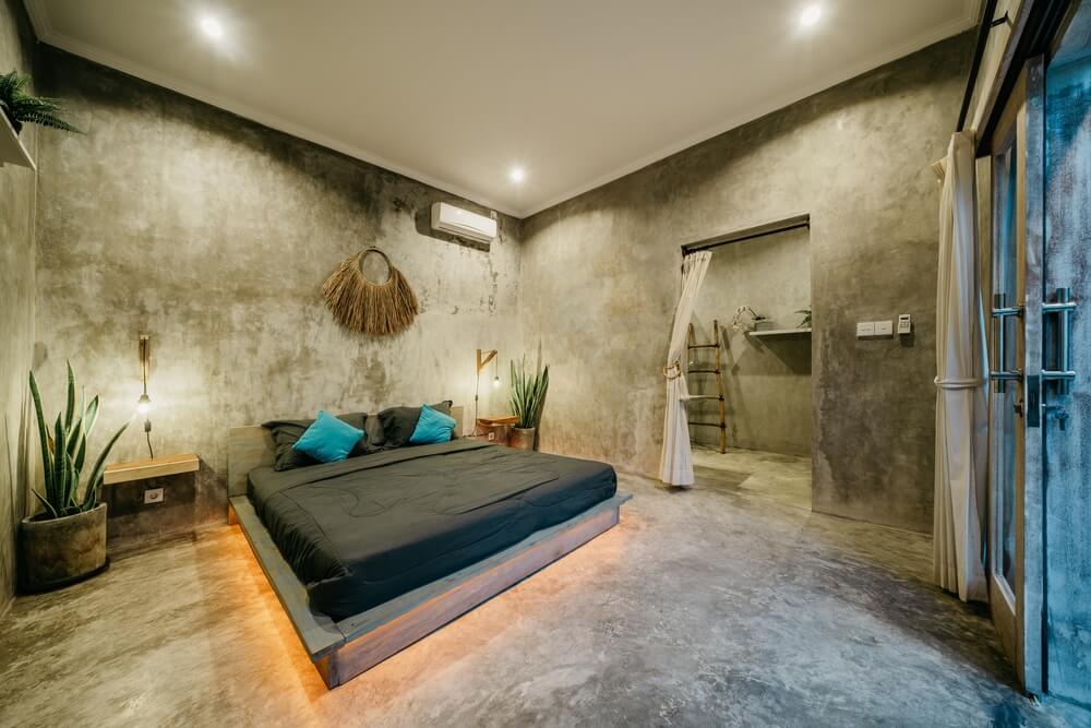 Quarto moderno e minimalista com paredes e piso de concreto, cama baixa de plataforma com roupa de cama preta, almofadas em tons de turquesa, plantas de interior, decoração natural e iluminação neon discreta embaixo da cama. Cortinas parcialmente abertas para portas de vidro.