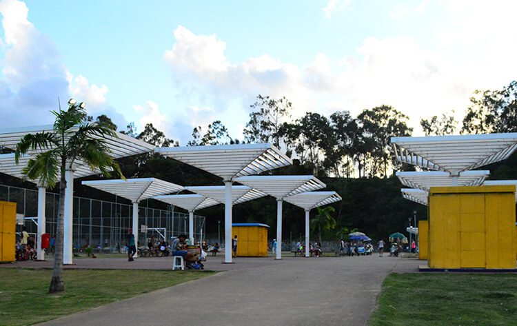 Uma área de lazer ao ar livre que lembra os parques do Recife, com modernas estruturas brancas de sombra, pessoas sentadas e caminhando, pequenos prédios amarelos, palmeiras, uma quadra esportiva e árvores sob um céu parcialmente nublado.