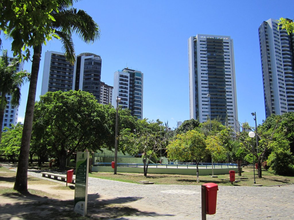 Um parque ensolarado com grandes árvores, uma passarela pavimentada, lixeiras vermelhas, placas informativas e altos prédios de apartamentos modernos ao fundo, sob um céu azul claro — um dos belos parques do Recife.