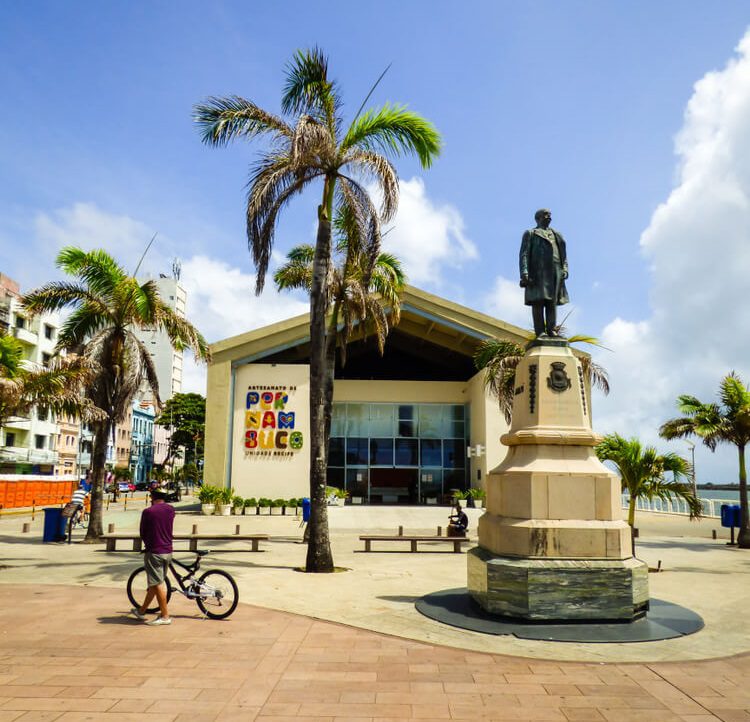 Uma pessoa com uma bicicleta está parada na Praça do Marco Zero, perto de uma estátua em um pedestal, com palmeiras e um prédio moderno ao fundo. O prédio tem uma placa colorida de Pernambuco na fachada.