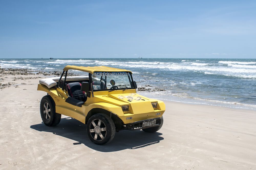 Um buggy amarelo está estacionado em uma praia de areia perto da costa nas Dunas Fortaleza, com o oceano e o céu azul ao fundo em um dia ensolarado.