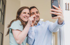 Foto que ilustra matéria sobre como montar uma carteira de investimentos para comprar um imóvel mostra um jovem casal tirando uma foto com o celular mostrando as chaves da casa nova