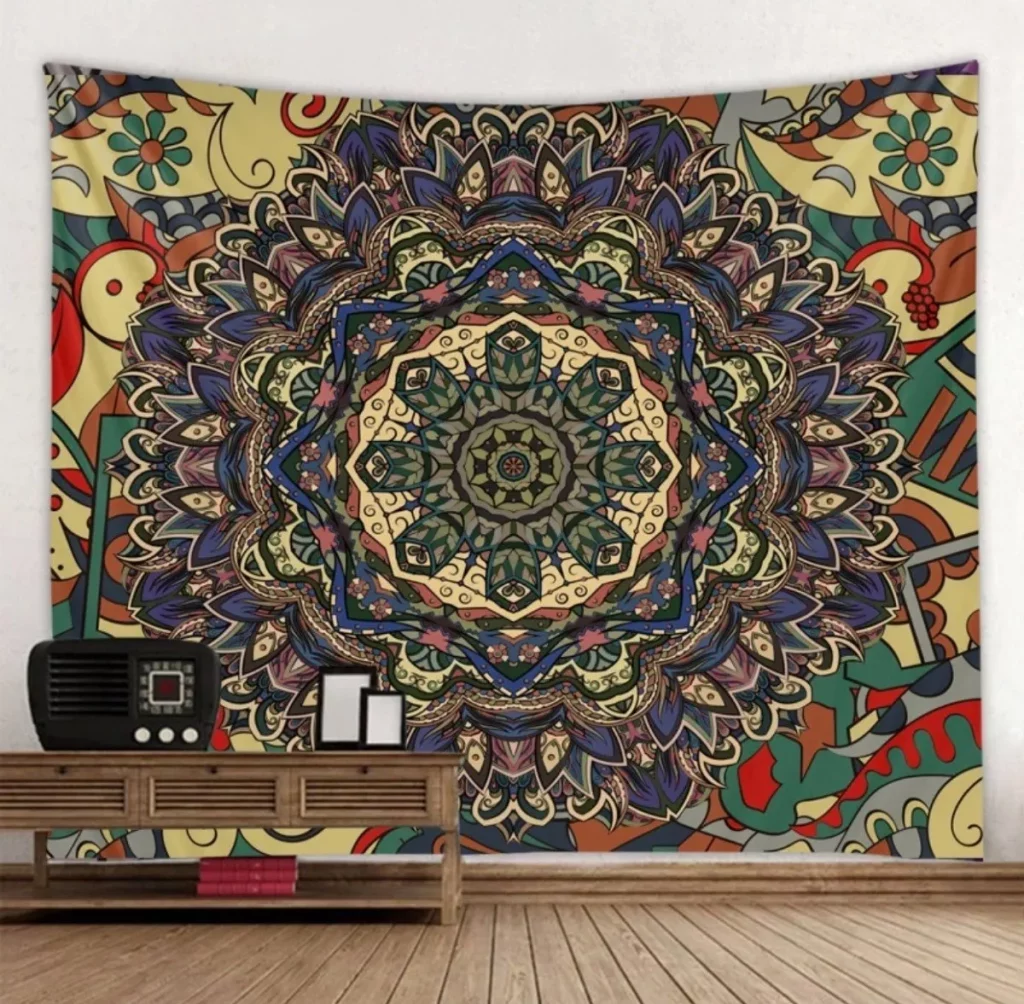 Uma tapeçaria de mandala colorida e simétrica com padrões florais e abstratos complexos está pendurada em uma parede de quarto decorada acima de uma mesa de madeira que contém um rádio antigo e uma moldura.