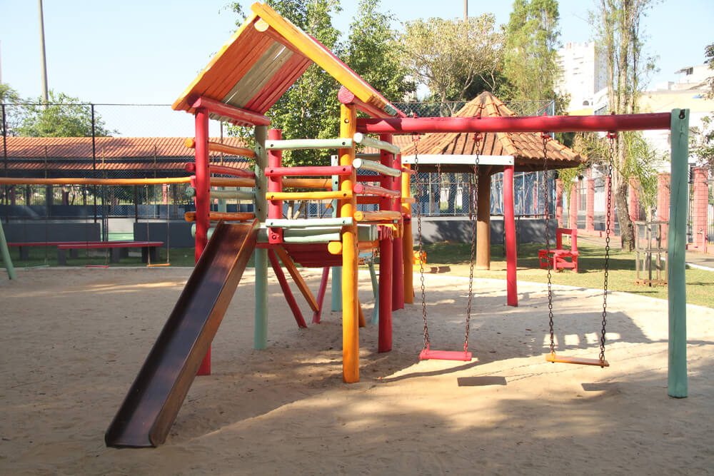 Um playground colorido no Parque Chico Mendes São Caetano conta com uma estrutura de escalada de madeira, um escorregador e dois balanços em terreno arenoso, todos cercados por árvores e uma cerca. A área ensolarada é vazia e convidativa.