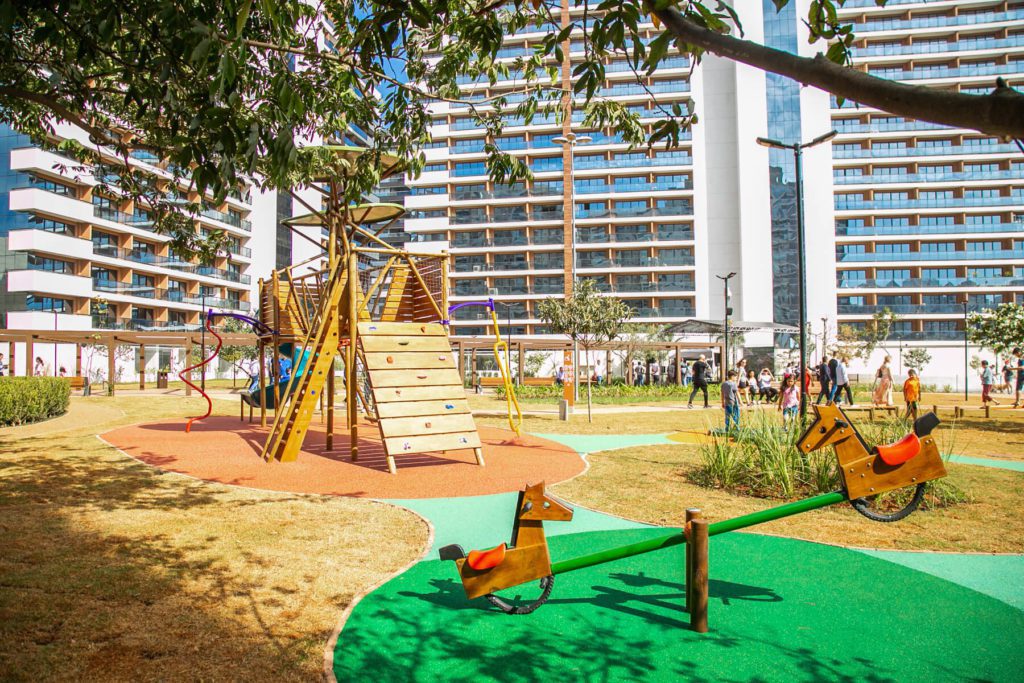 Um playground moderno com estruturas de escalada, escorregadores e gangorras fica sobre um piso de borracha colorido no Parque Chico Mendes São Caetano, cercado por grama e árvores, com altos prédios de apartamentos e pessoas ao fundo em um dia ensolarado.