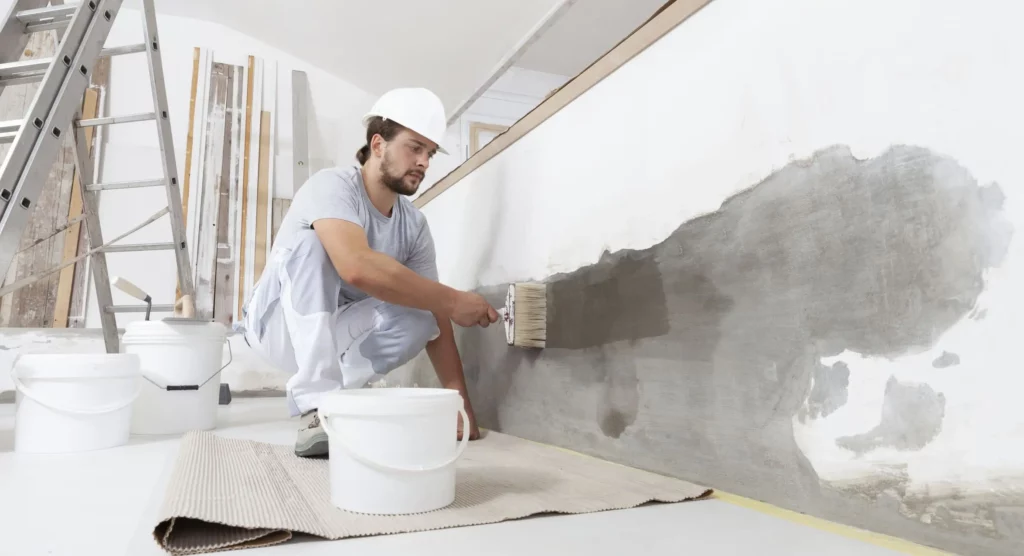 Imagem de um trabalhador da construção civil, usando capacete e macacão, ajoelhado em um tapete enquanto pinta uma grande parede interna. Um balde branco está próximo