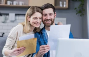 Imagem de um casal sorridente segurando e olhando para documentos e um envelope, parecendo feliz e animado. Eles estão em um ambiente interno, com uma cozinha visível ao fundo