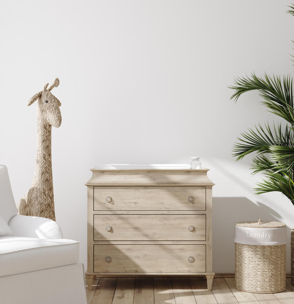 Um aconchegante quarto de bebê simples conta com uma poltrona branca, uma girafa de vime, uma cômoda de madeira clara, um cesto de roupa suja e uma planta verde em vaso contra uma parede branca.