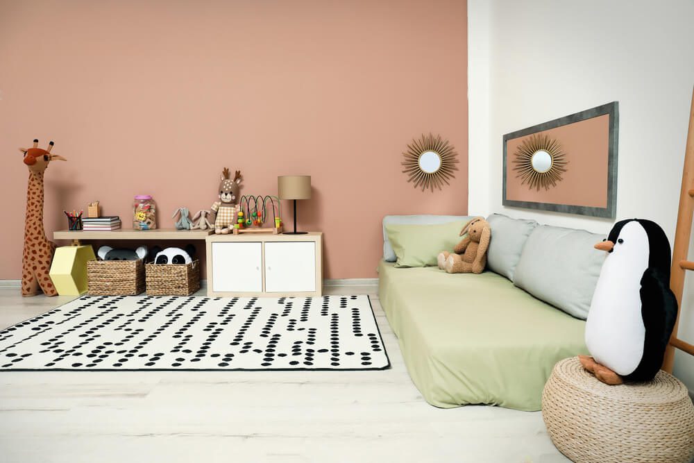 Um quarto aconchegante para bebê simples com uma girafa de brinquedo, um coelho de pelúcia e um pinguim de pelúcia, uma cama verde macia, prateleiras com cestos e brinquedos, espelhos de parede e um tapete branco com bolinhas pretas sobre um piso de madeira clara.