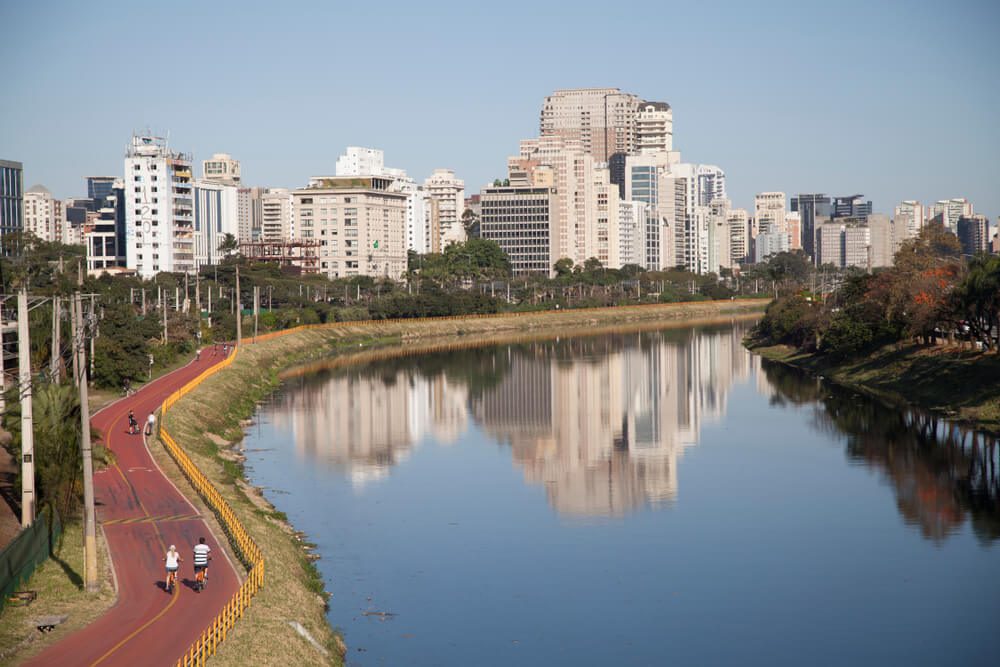 Uma ciclovia e ciclovia à beira do rio, com pessoas usando-a, ladeada por árvores verdes de um lado e altos edifícios modernos do outro, refletidos nas águas calmas sob um céu azul claro.
