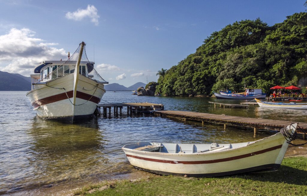 Imagem de um grande barco branco atracado em um píer de madeira em um lago calmo na Lagoa da Conceição, em Florianópolis, com um pequeno barco a remo na margem gramada, para ilustrar matéria sobre o que fazer em Florianópolis em casal