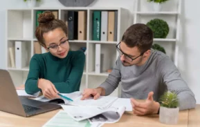 Imagem de um homem e uma mulher sentados um ao lado do outro em uma mesa analisando informações contidas em livros e outros documentos em cima da mesa de madeira e ao lado de um notebook para ilustrar matéria sobre juros de mora