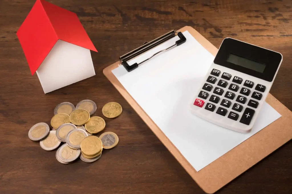 Imagem de uma caderneta, moedas, uma calculadora e uma casa de madeira em tamanho miniatura em cima de uma mesa de madeira para ilustrar matéria sobre juros moratórios