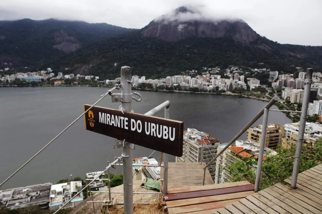 Imagem de uma placa com a inscrição “Mirante do Urubu” tem vista para uma paisagem urbana com prédios, um corpo d’água e, ao fundo, uma montanha envolta em nuvens no Parque da Catacumba, no Rio de Janeiro