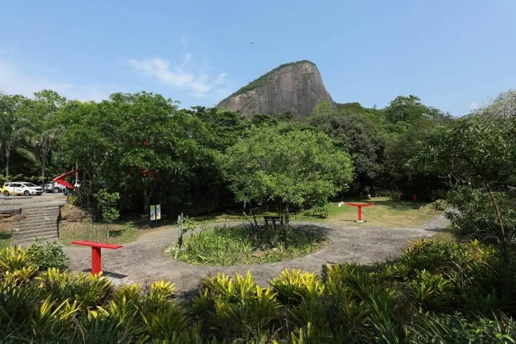 Imagem da vista do Parque Natural Municipal Penhasco Dois Irmãos, no Rio de Janeiro, mostra um jardim com um caminho circular, esculturas vermelhas, árvores e uma colina rochosa ao fundo