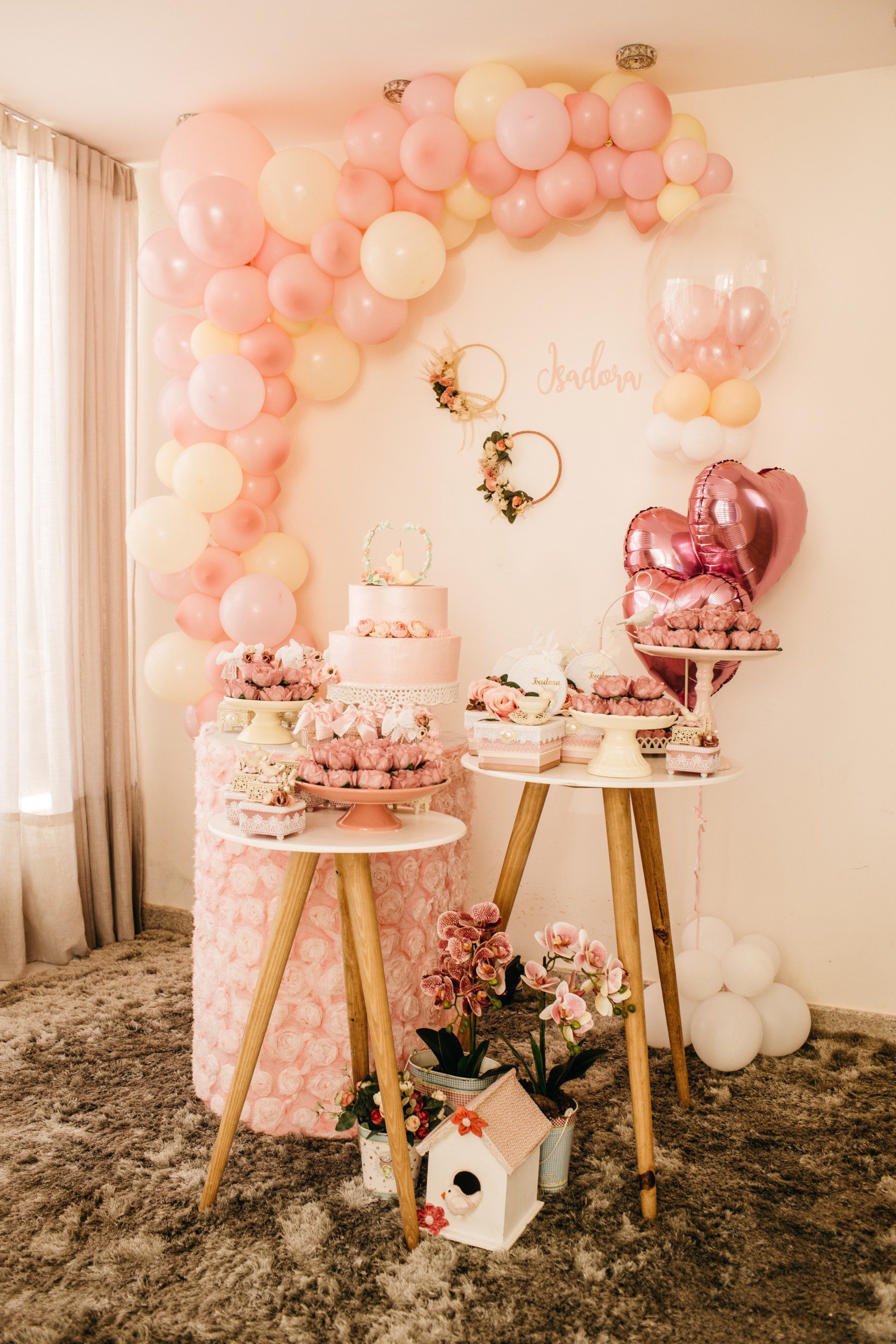 Imagem de uma decoração de festa simples em tons de rosa com arco de balões desconstruído, quadrinhos na parede, arranjos de flores e bolo de andares.