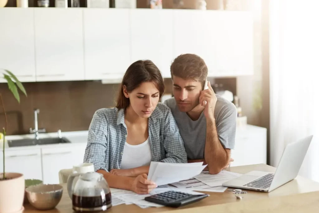 Imagem de um casal formado por um homem e uma mulher sentados analisando informações em papéis que estão em cima de uma mesa ao lado de um notebook, chaves, calculadora e uma garrafa de café para ilustrar matéria sobre taxa de juros de mora