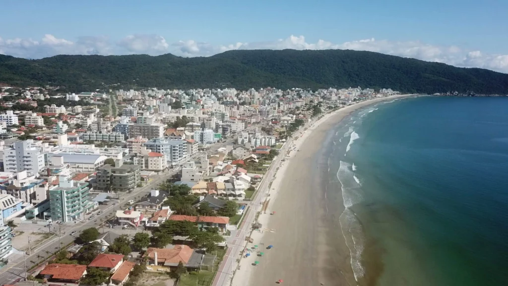 Imagem da vista aérea do litoral de Bombinhas mostra mar, faixa de areia, avenidas, casas, prédios e montanhas ao fundo para ilustrar matéria sobre as melhores cidades de Santa Catarina