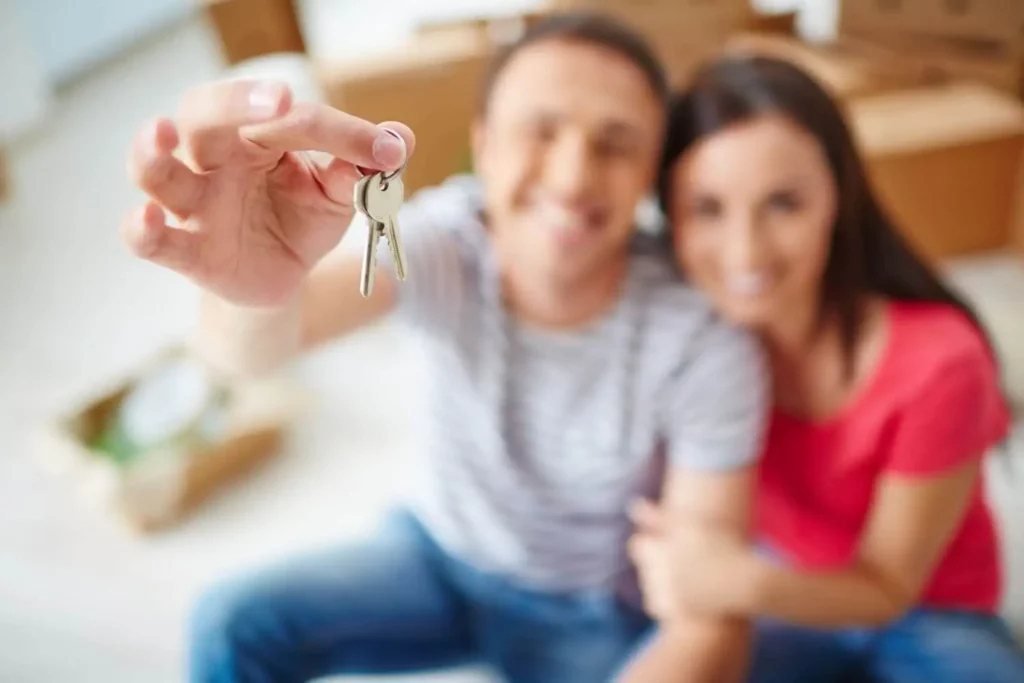 Imagem de um homem e uma mulher sentados no chão de um imóvel na frente de algumas caixas de papelão segurando as chaves de casa para ilustrar matéria sobre o que é pagar caução de aluguel