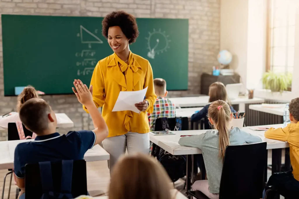 Imagem de uma professora tocando na mão de um aluno enquanto anda pela sala de aula com uma folha de papel na mão para ilustrar matéria sobre escolas em Osasco, particulares