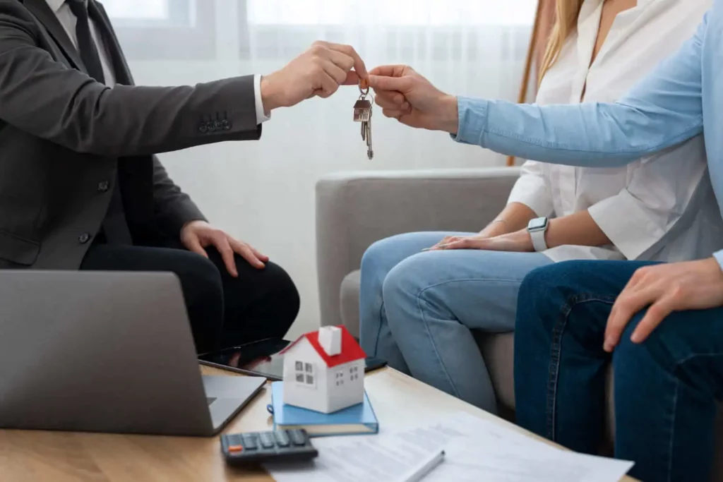 Imagem de um agente imobiliário entregando as chaves de um imóvel para um homem que está sentado ao lado de uma mulher em um sofá em frente a uma mesinha de centro com um notebook, papéis, calculadora e uma casa de madeira em tamanho miniatura em cima para ilustrar matéria sobre o que é caução no aluguel de imóvel