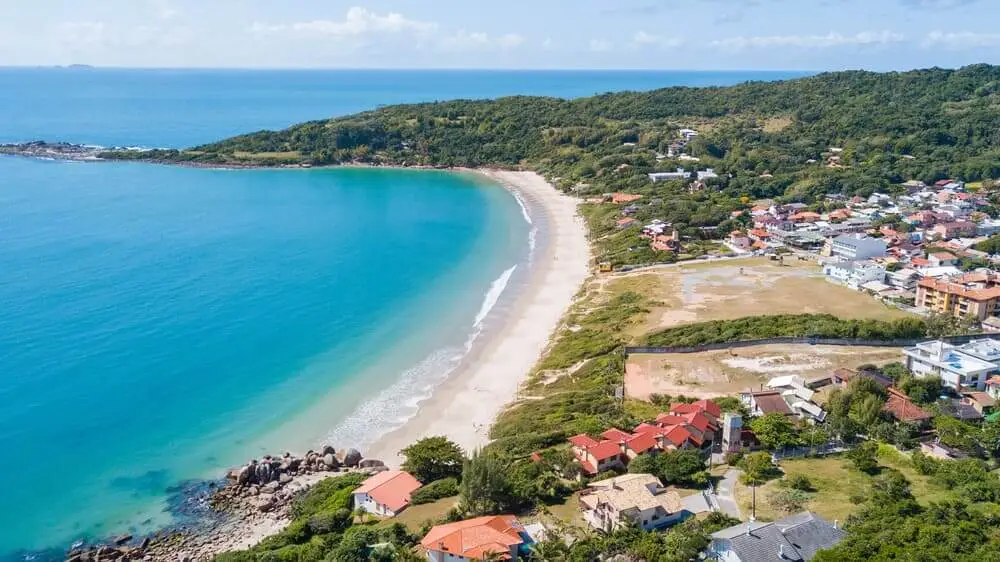 Imagem da vista aérea das Praias de Palhoça, mostrando uma praia em formato de meia-lua com águas azul-turquesa, cercada por morros verdes e casas ao longo da costa sob um céu azul com nuvens dispersas, para ilustra matéria sobre as melhores cidades para viver em Santa Catarina