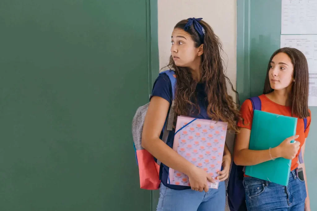 Imagem de duas estudantes adolescentes encostadas na parede olhando para o lado enquanto seguram o material escolar para ilustrar matéria sobre quantas escolas têm em Osasco